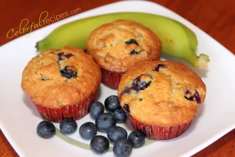Three moist banana blueberry muffins on a white plate with fresh blueberries and a banana