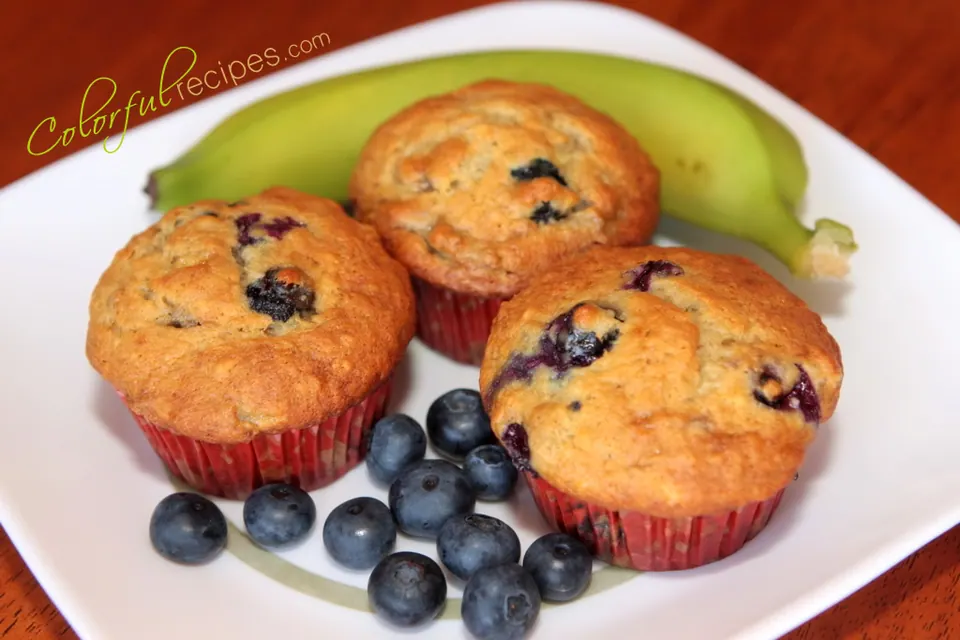 Three banana blueberry muffins on a white plate with fresh blueberries and a banana
