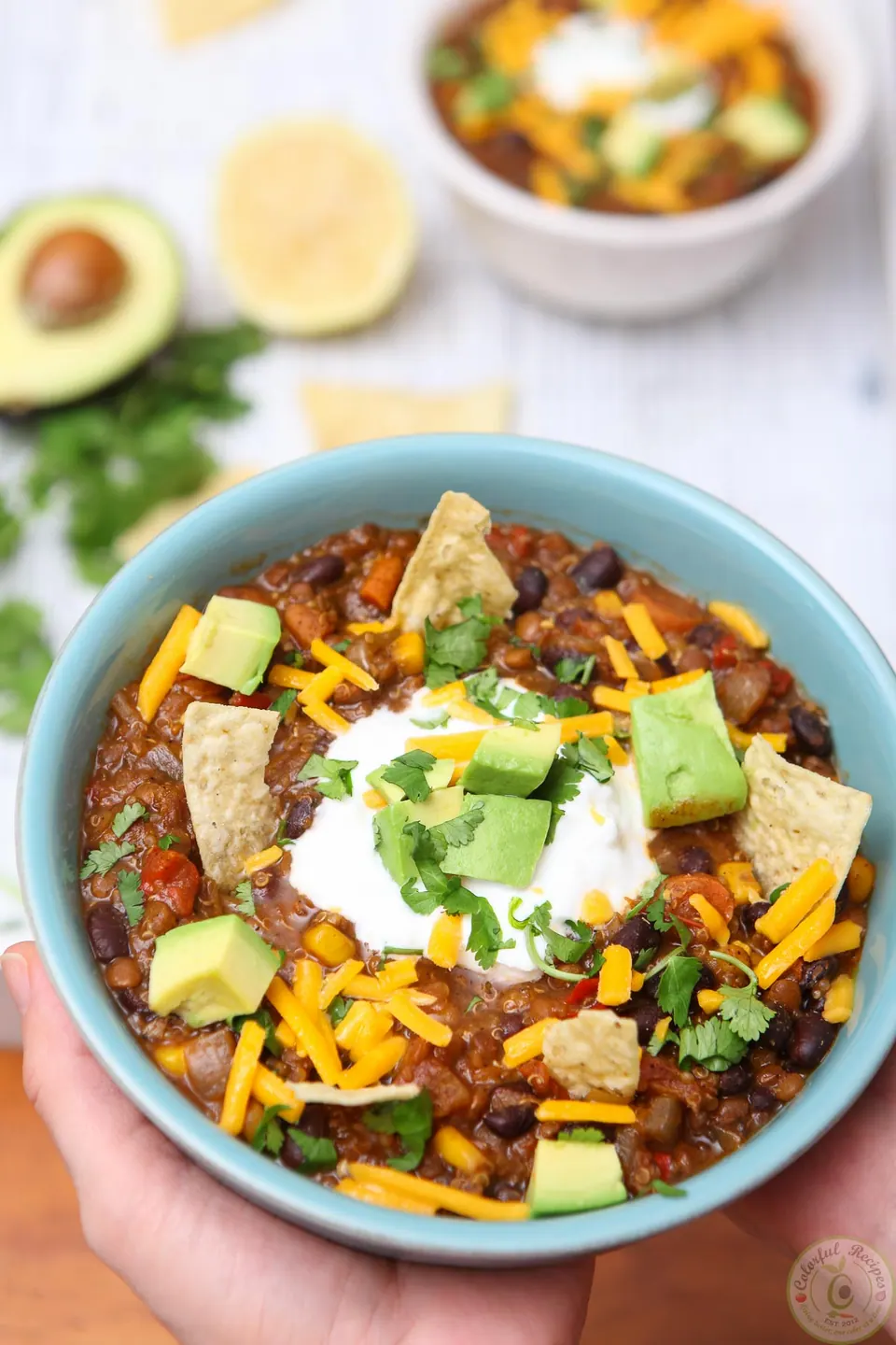 Hands holding a bowl of black bean lentil quinoa chili topped with avocado, cheddar and chips