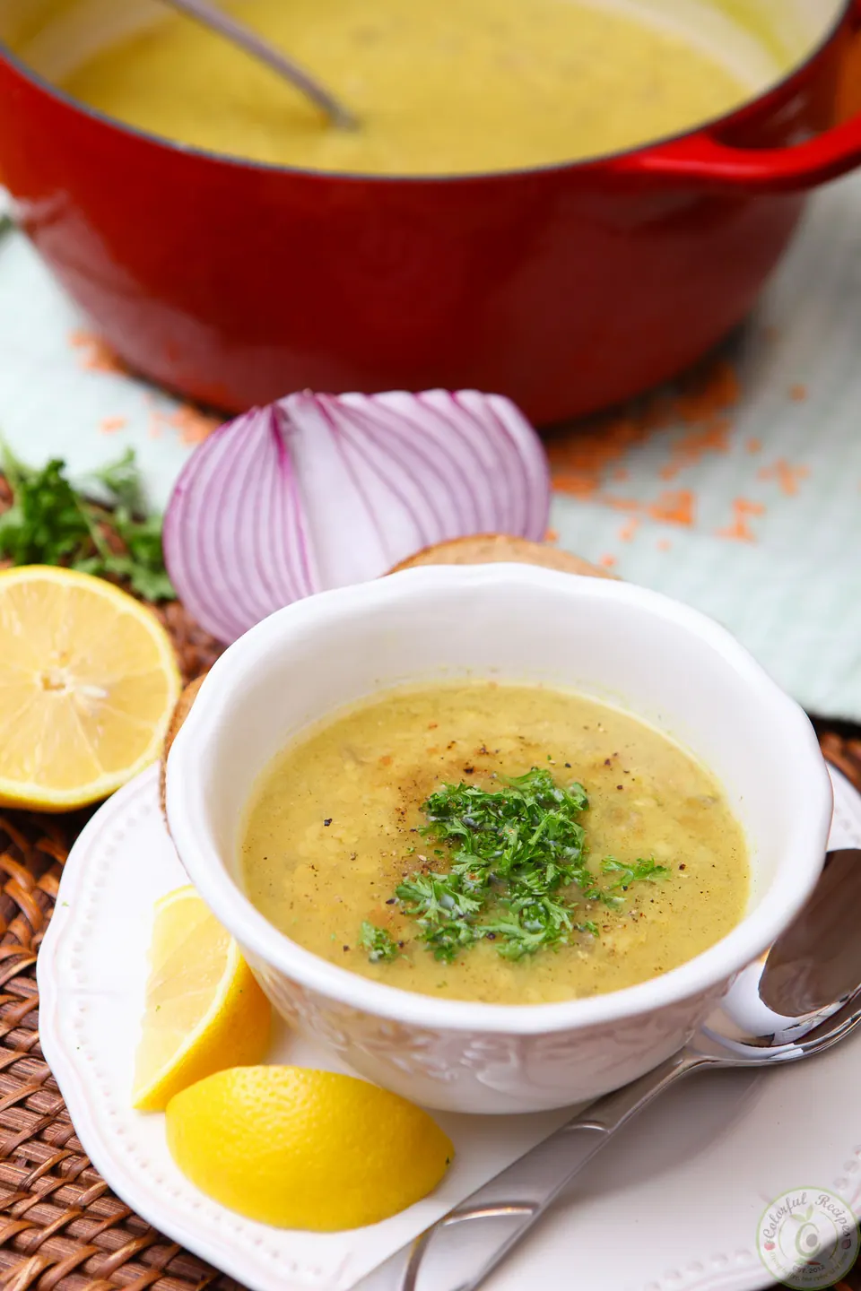 Bowl of Shorbet Adas Mediterranean red lentil soup with parsley, lemon wedges and a pot behind it