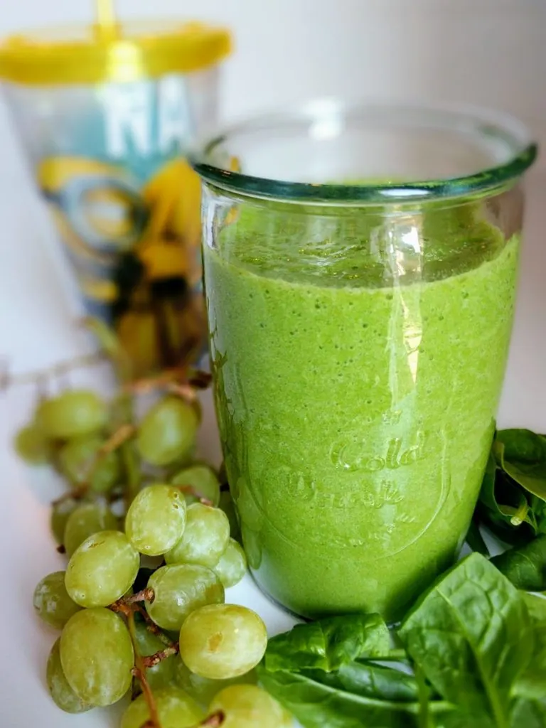 Glass of banana pineapple green smoothie next to grapes and spinach leaves