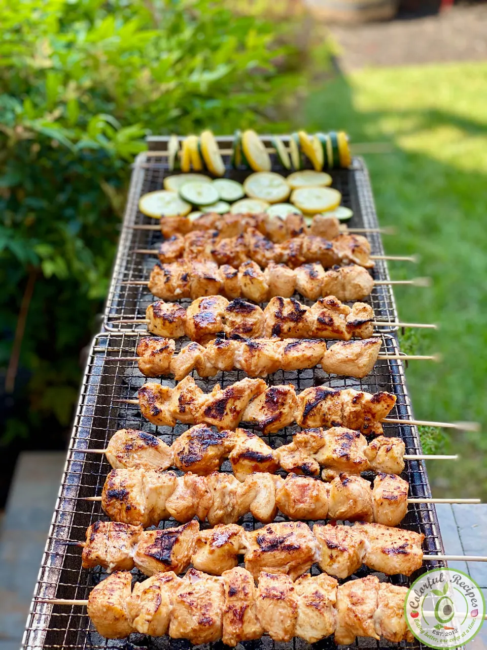 Chicken kabobs and sliced zucchini and squash cooking on an open mankal grill