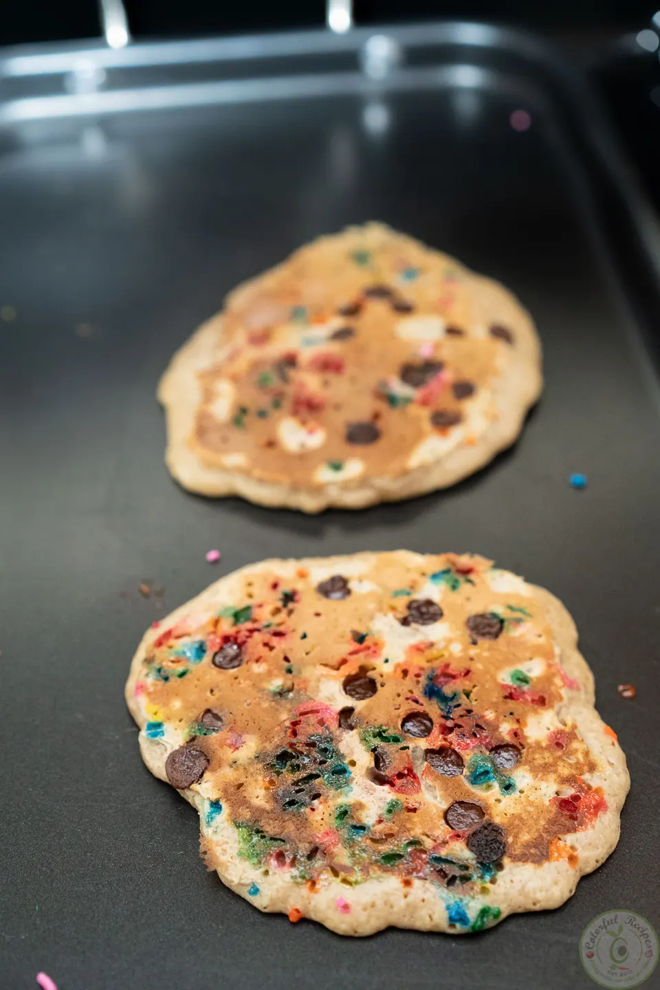 Two sprinkle and chocolate chip oatmeal pancakes on a griddle