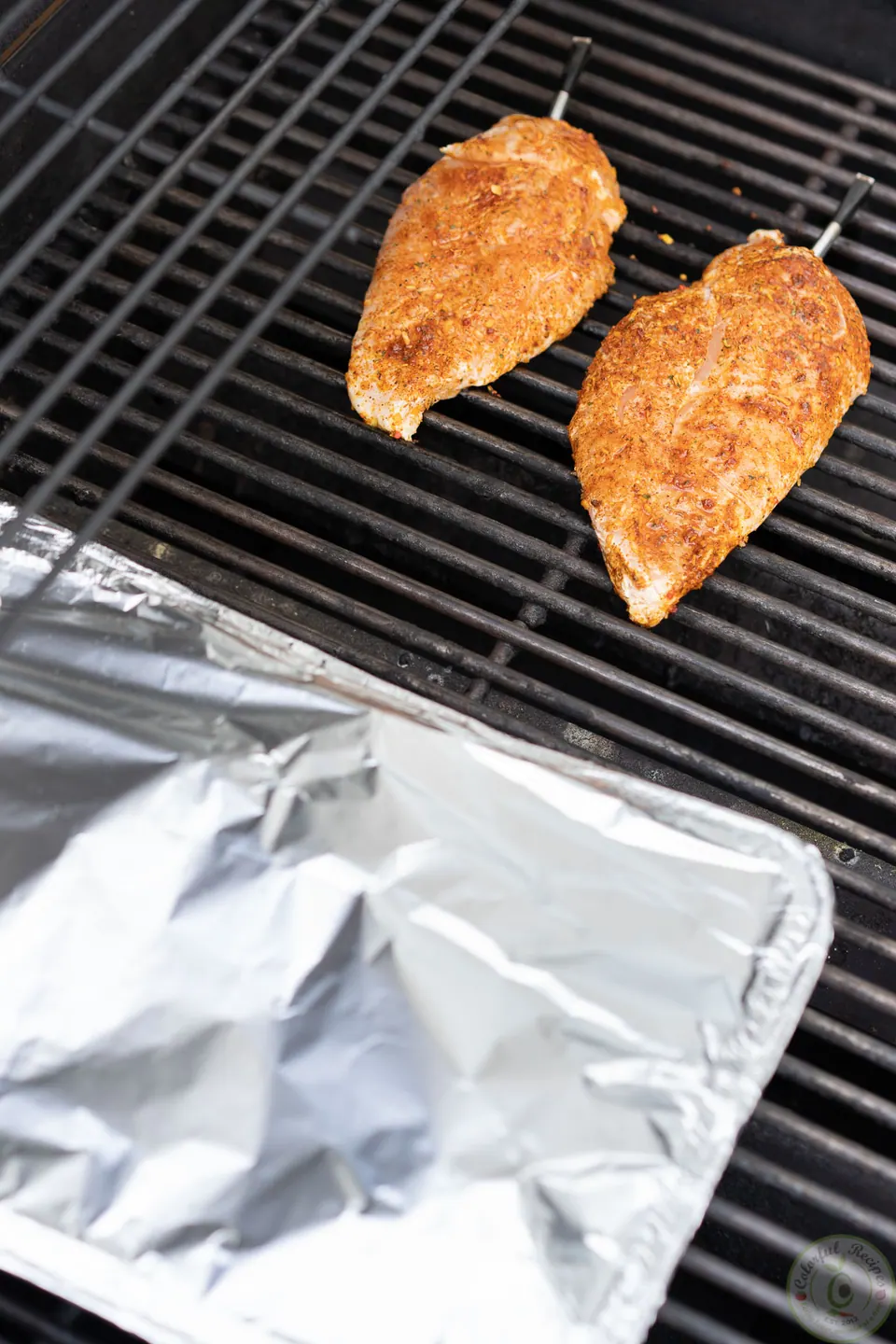 Seasoned chicken breasts on the grill next to a foil-covered tray of veggies