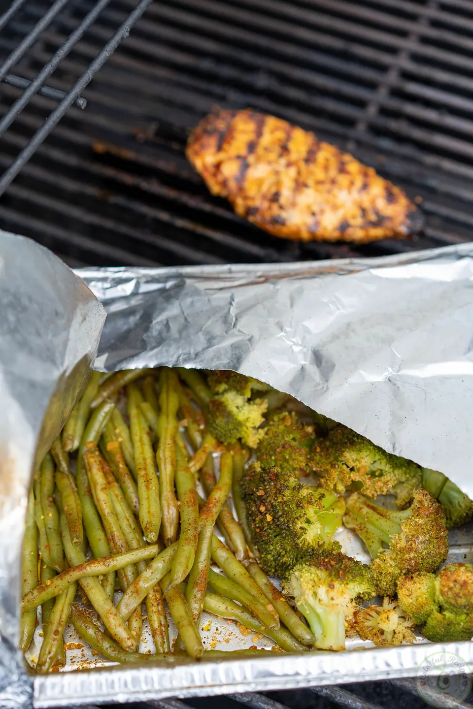 Grilled chicken breast beside a foil tray of steamed green beans and broccoli