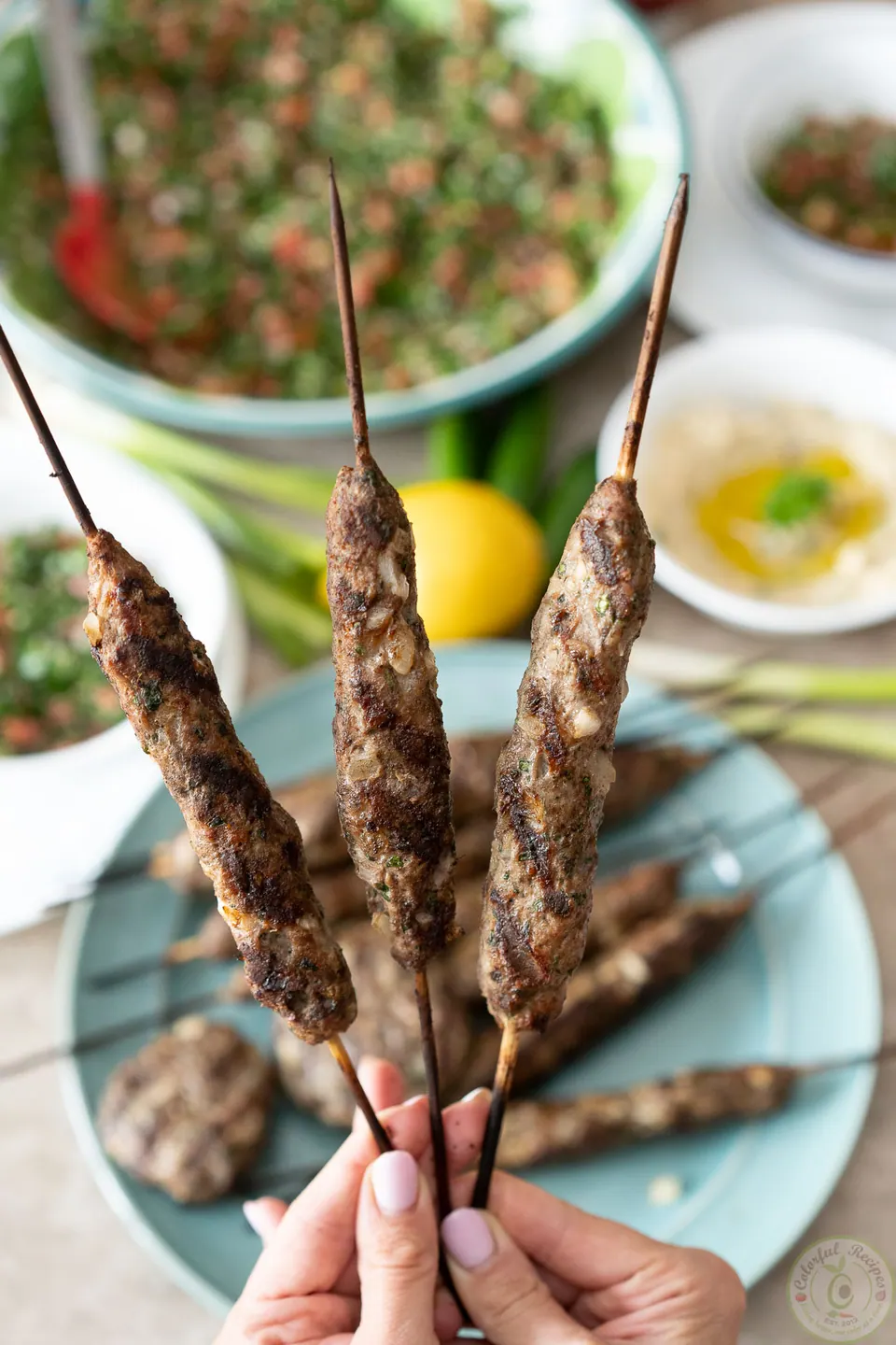 Hands holding grilled lamb kafta kabob skewers over a plate