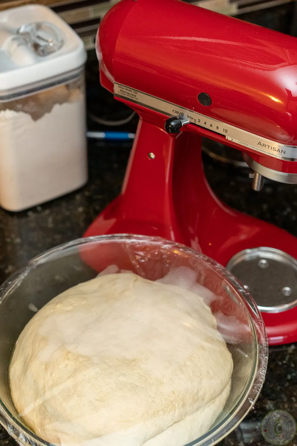 Risen pizza dough doubled in size in a glass bowl next to a stand mixer