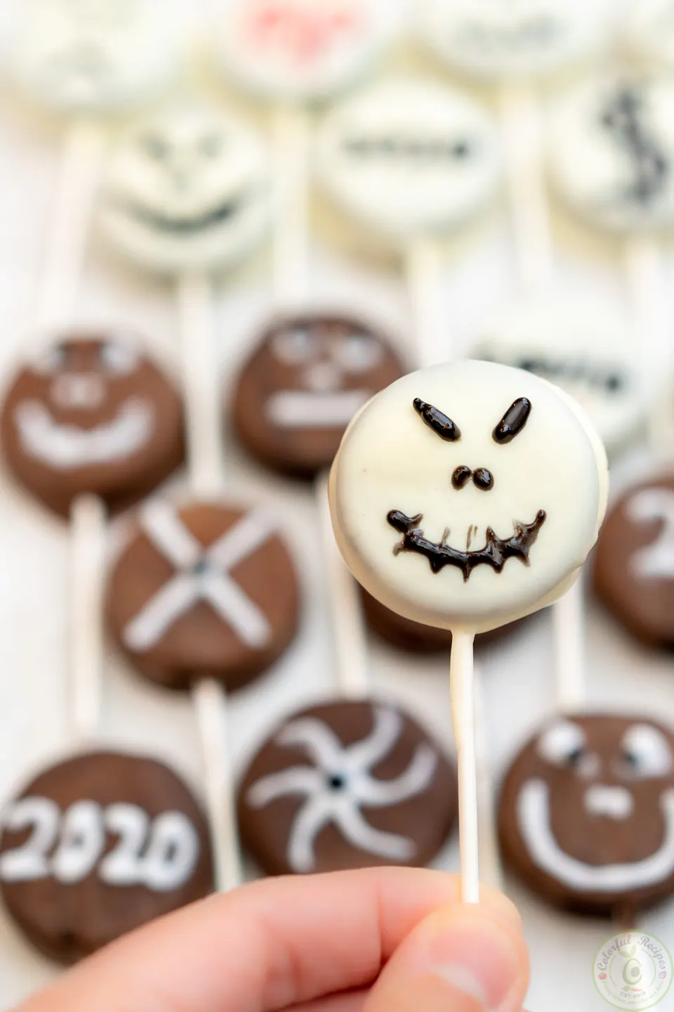 Hand holding a white chocolate Halloween Oreo pop with a jack-o’-lantern face